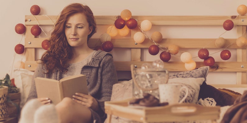 May include: A woman sits in bed reading a book. The bed is decorated with string lights and pillows. There is a tray with a mug and a glass jar on a small table next to the bed.
