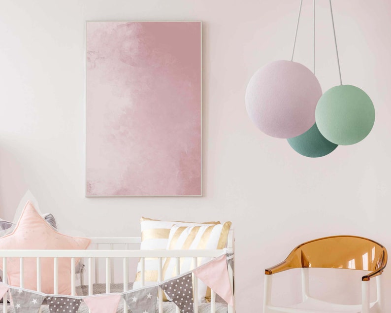 May include: A white crib with a pink and gray patterned crib bumper and a pink and gray flag banner. A pink and gold pillow is on the crib mattress. A pink, green, and white hanging light fixture is in the background. A clear plastic chair is in the foreground.
