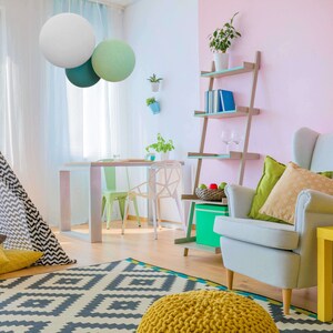 May include: A living room with a white, gray, and green lampshade hanging from the ceiling. There is a teepee tent, a yellow and gray rug, a yellow ottoman, a white armchair with pillows, a yellow side table, and a bookshelf with books and glasses.