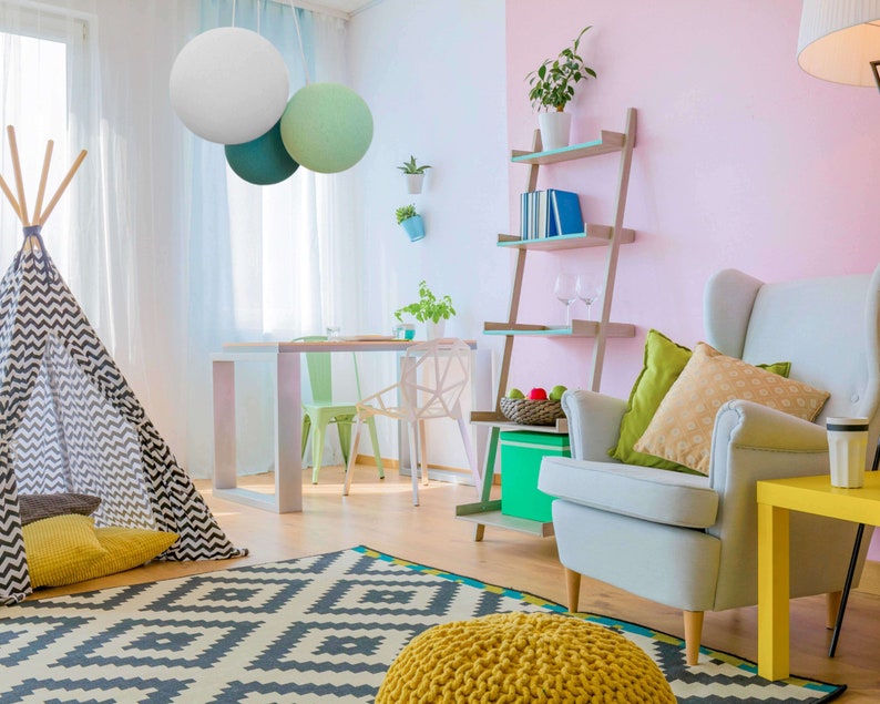 May include: A living room with a white, gray, and green lampshade hanging from the ceiling. There is a teepee tent, a yellow and gray rug, a yellow ottoman, a white armchair with pillows, a yellow side table, and a bookshelf with books and glasses.