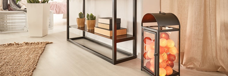 May include: A black metal lantern with a glass panel filled with orange and white cotton ball lights. The lantern is sitting on a wooden floor in front of a beige curtain.