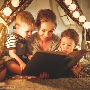 May include: A woman reads a book to two children inside a tent decorated with white string lights. The children are sitting on a soft, plush rug.