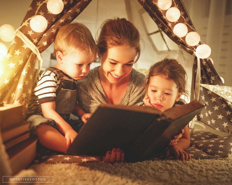 May include: A woman reads a book to two children inside a tent decorated with white string lights. The children are sitting on a soft, plush rug.
