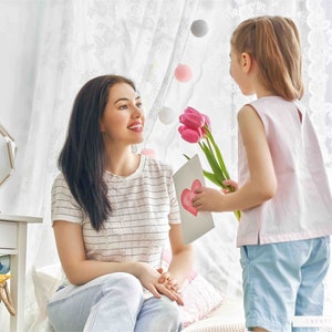 May include: A young girl gives her mother a card and pink flowers. The card has a heart drawn on it. The girl is wearing a pink sleeveless shirt and blue shorts. The mother is wearing a striped shirt and blue jeans.
