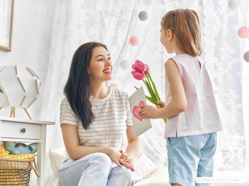 May include: A young girl gives her mother a card and pink flowers. The card has a heart drawn on it. The girl is wearing a pink sleeveless shirt and blue shorts. The mother is wearing a striped shirt and blue jeans.