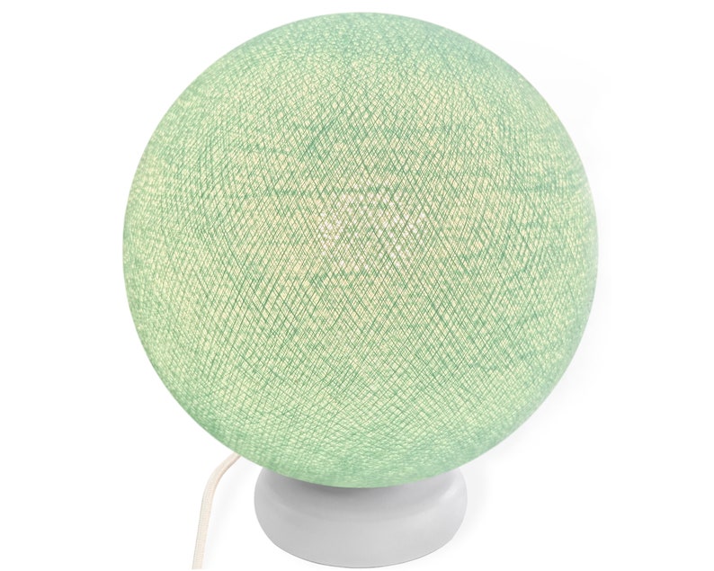 Pode incluir: Um abajur de mesa de bola de algod&atilde;o verde com uma base branca. A l&acirc;mpada est&aacute; acesa e emite um brilho quente.