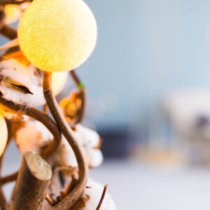 May include: A string of cotton ball lights wrapped around a branch with white cotton balls. The lights are a soft yellow color and the branch is brown.