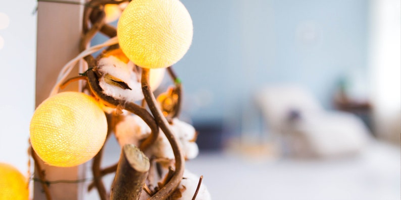 May include: A string of cotton ball lights wrapped around a branch with white cotton balls. The lights are a soft yellow color and the branch is brown.