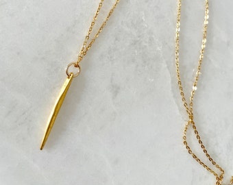May include: A gold chain necklace with a long, thin, gold pendant. The pendant is shaped like a sharp, pointed blade.