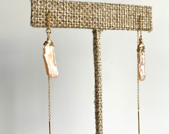 May include: A pair of gold-colored threader earrings. Each earring features a long, slender, light pink baroque pearl wrapped in gold wire, suspended from a delicate chain. A gold bar hangs at the end of each chain.
