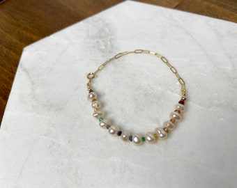 May include: A delicate gold-toned bracelet featuring a chain link design and a strand of white pearls. The bracelet is accented with small, colorful gemstones, including green, blue, and red. The jewelry is displayed on a white marble surface.
