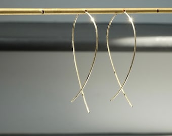 May include: A pair of gold-colored, curved earrings. The earrings have a delicate, elongated design, with a slender wire that forms a looping shape. The earrings are displayed against a neutral background, highlighting their simple elegance.