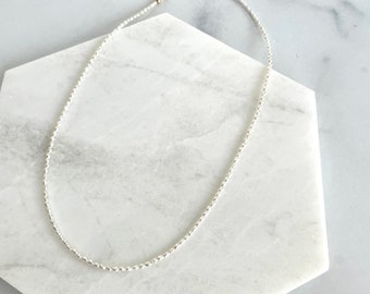 May include: A delicate necklace composed of numerous small, reflective silver-toned beads strung together in a continuous, uniform chain. The beads have a subtle, textured surface that catches the light, creating a shimmering effect along the entire length of the strand.