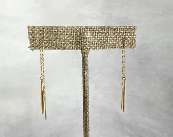 May include: A pair of gold-colored threader earrings. Each earring features a delicate chain that extends through the ear, with a slender, elongated charm suspended at the end. The earrings are displayed on a neutral-toned jewelry stand.