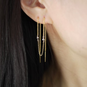 May include: Gold chain earrings with small silver beads hanging from a gold bar. The earrings are worn in a person's ear.