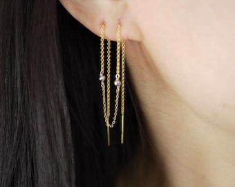 May include: Gold chain earrings with small silver beads hanging from a gold bar. The earrings are worn in a person's ear.