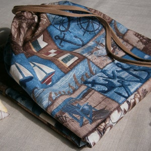 May include: A blue and brown fabric drawstring bag with a nautical theme. The bag features anchors, ships, and other nautical imagery. The bag has a brown drawstring and a brown wooden bead.