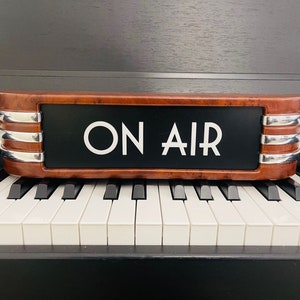 Op de afbeelding: Een rechthoekig "ON AIR" bord met een bruin houten frame en zilveren accenten, rustend op een pianoklavier. Het bord heeft een zwarte achtergrond met witte letters. De pianotoetsen zijn zwart en wit.