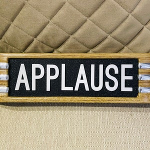 以下が含まれることがあります： 白い大文字で「APPLAUSE」と書かれた長方形の木製サイン。サインは黒い背景で、明るい木材と銀色の金属トリムで縁取られています。サインはベージュの表面に置かれています。