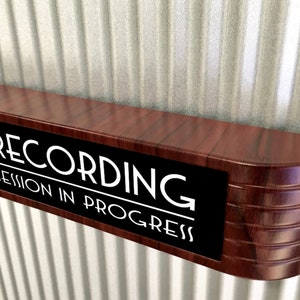 Può includere: Un cartello in legno con le parole "RECORDING SESSION IN PROGRESS" in lettere bianche su sfondo nero. Il cartello ha una forma curva ed è montato su una superficie di metallo ondulato.
