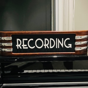 May include: A rectangular wooden sign with the word "RECORDING" in white lettering. The sign has a black background and is framed with brown wood and silver accents. It sits atop a black piano.