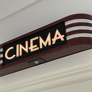May include: A rectangular "CINEMA" sign with a dark wood-grain finish and silver accents. The illuminated letters are a warm, golden color. The sign has a small black sensor on the bottom.