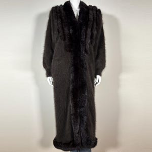 May include: A long, black, fuzzy cardigan with a deep V-neck and a contrasting black trim. The cardigan is made of a soft, luxurious fabric and has a relaxed fit.