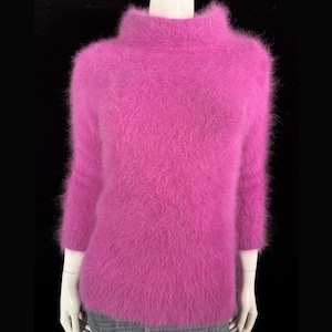 May include: A vibrant magenta sweater with a mock turtleneck and three-quarter sleeves. The fuzzy knitwear is displayed on a mannequin against a black background. The sweater's texture appears soft and plush.