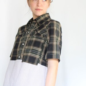May include: A woman wearing a short-sleeved plaid shirt with a white striped shirt underneath. The plaid shirt is black, green, and tan.
