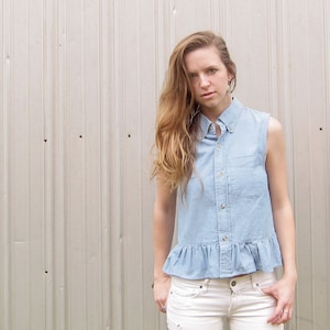 May include: A woman wearing a light blue denim shirt with a ruffled hem and white jeans. The shirt has a collared neckline and button closure.