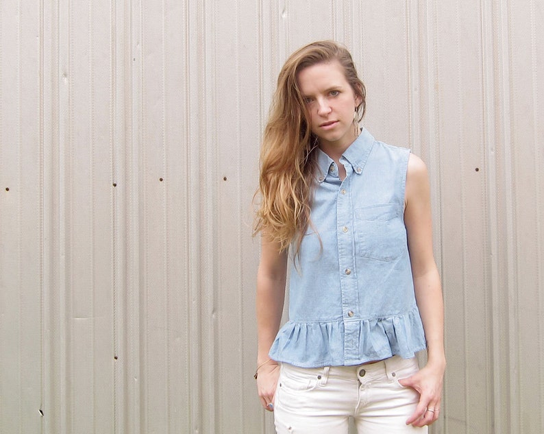 May include: A woman wearing a light blue denim shirt with a ruffled hem and white jeans. The shirt has a collared neckline and button closure.