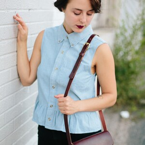 May include: A woman wearing a light blue denim shirt with a brown leather shoulder bag. The bag has a gold buckle and a flap closure.