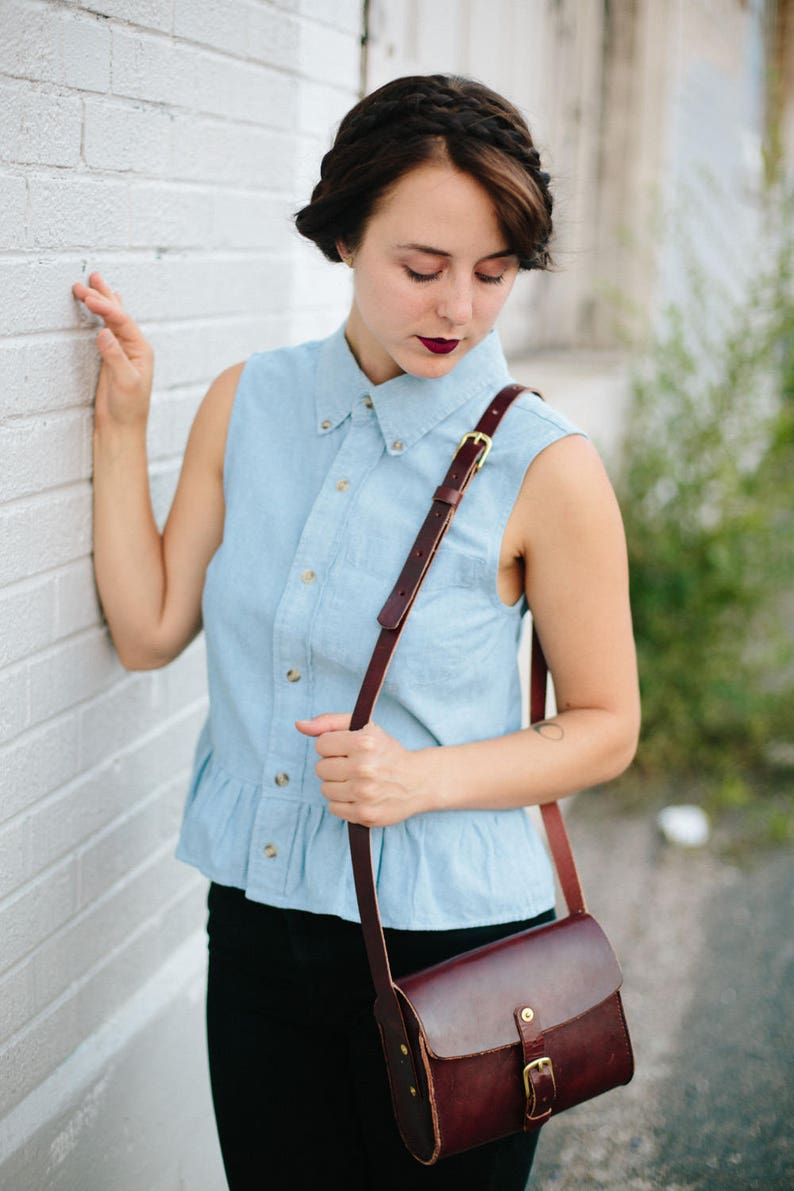 May include: A woman wearing a light blue denim shirt with a brown leather shoulder bag. The bag has a gold buckle and a flap closure.