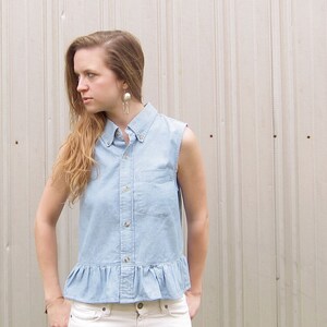 May include: A light blue denim shirt with a ruffled hem and a button-up front. The shirt has short sleeves and a collared neckline.