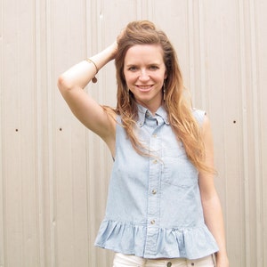 May include: A woman wearing a light blue denim shirt with a frilled hem and a collared neckline. She is standing in front of a white wall.