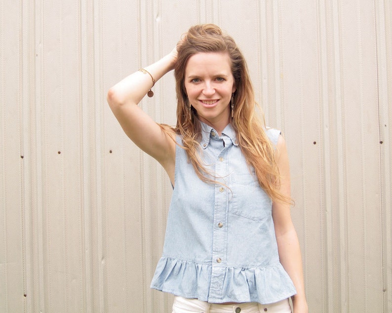 May include: A woman wearing a light blue denim shirt with a frilled hem and a collared neckline. She is standing in front of a white wall.
