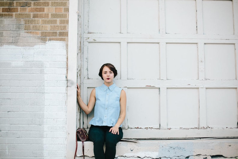 May include: A woman wearing a light blue collared shirt with a frilled hem, black trousers, and a brown leather bag sits in front of a white garage door.