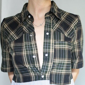 May include: A green and black plaid shirt with a white striped bottom. The shirt is unbuttoned and has a silver cross necklace visible.