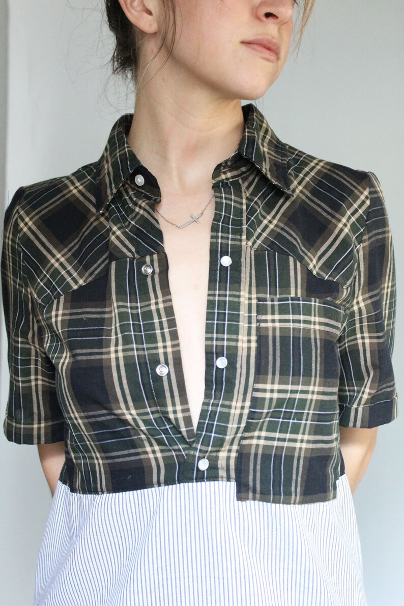 May include: A green and black plaid shirt with a white striped bottom. The shirt is unbuttoned and has a silver cross necklace visible.