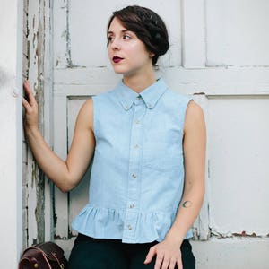 May include: A woman wearing a light blue sleeveless button-down shirt with a ruffled hem, black trousers, and a brown leather satchel bag. She has dark hair styled in a braid and is looking to the left.