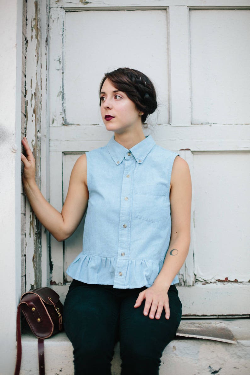May include: A woman wearing a light blue sleeveless button-down shirt with a ruffled hem, black trousers, and a brown leather satchel bag. She has dark hair styled in a braid and is looking to the left.