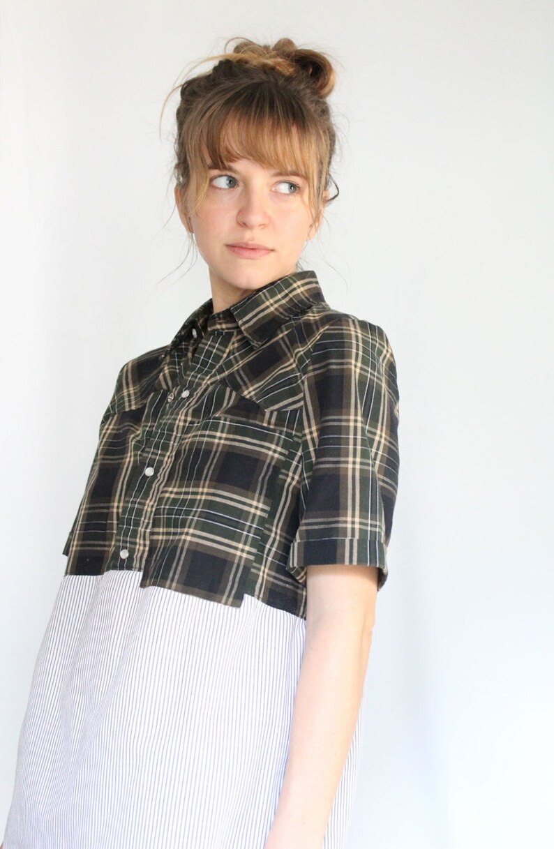 May include: A woman wearing a short-sleeved plaid shirt with a white striped shirt underneath. The plaid shirt is black, green, and tan.