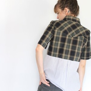 May include: A woman wearing a black and green plaid shirt with a white striped shirt underneath. The plaid shirt is cropped and the striped shirt is longer. She is wearing gray jeans.