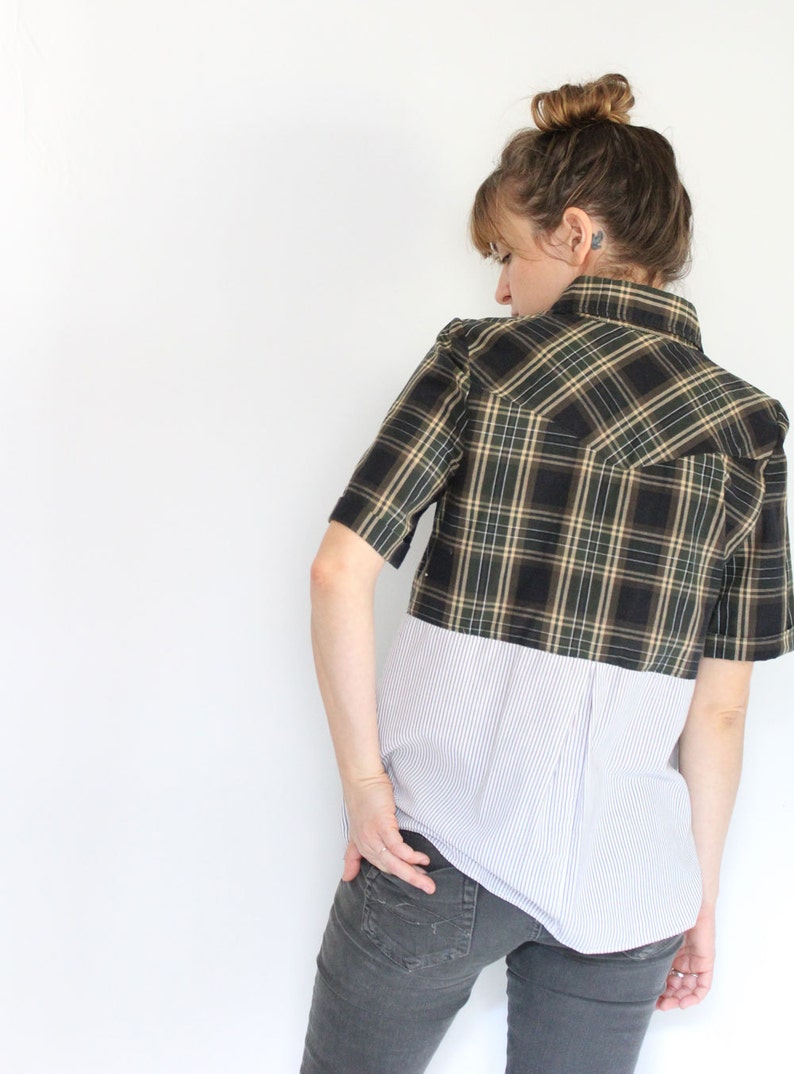 May include: A woman wearing a black and green plaid shirt with a white striped shirt underneath. The plaid shirt is cropped and the striped shirt is longer. She is wearing gray jeans.