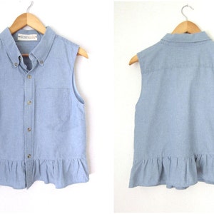 May include: A light blue sleeveless denim shirt with a frilled hem and a button-up front. The shirt has a small pocket on the left side and a label that says "Honeycomb".