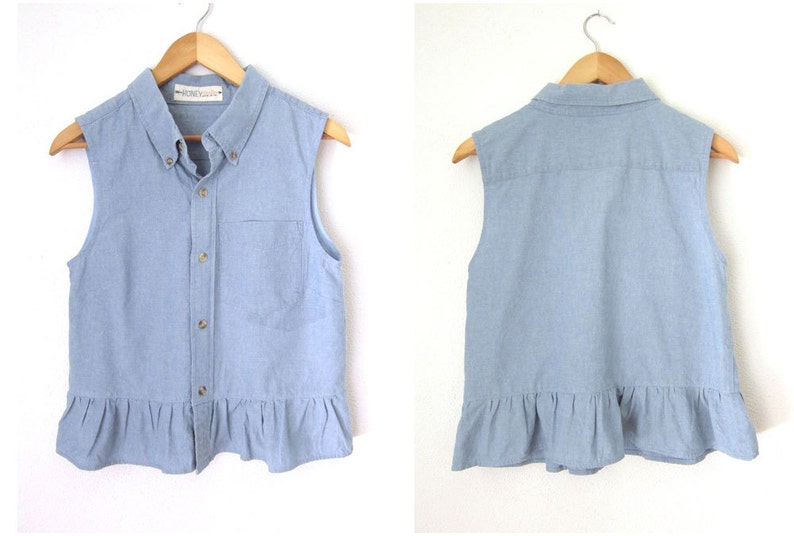 May include: A light blue sleeveless denim shirt with a frilled hem and a button-up front. The shirt has a small pocket on the left side and a label that says "Honeycomb".