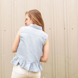 May include: A light blue denim shirt with a ruffled hem and a collared neckline. The shirt is sleeveless and has a casual, relaxed fit.