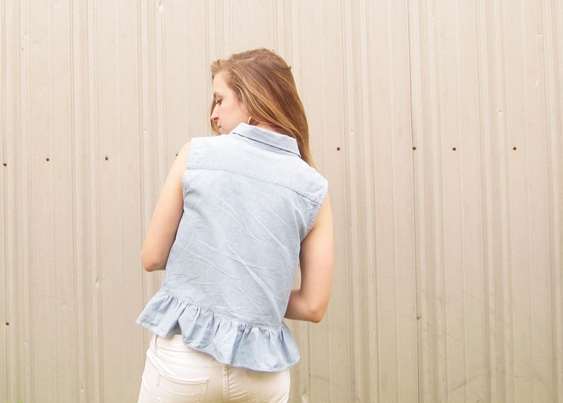 May include: A light blue denim shirt with a ruffled hem and a collared neckline. The shirt is sleeveless and has a casual, relaxed fit.