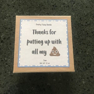May include: A brown box with a white label that says "Pretty Fizzy Bombs" and "Thanks for putting up with all my" with a cartoon poop emoji. The label also says "Soap" and "Net. Wt. 1.2 oz".