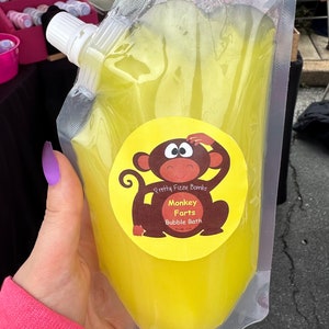 May include: A yellow pouch filled with a yellow liquid. The pouch has a label that says "Pretty Fizzy Bombs" and "Monkey Farts Bubble Bath". A cartoon monkey with big eyes is on the label.